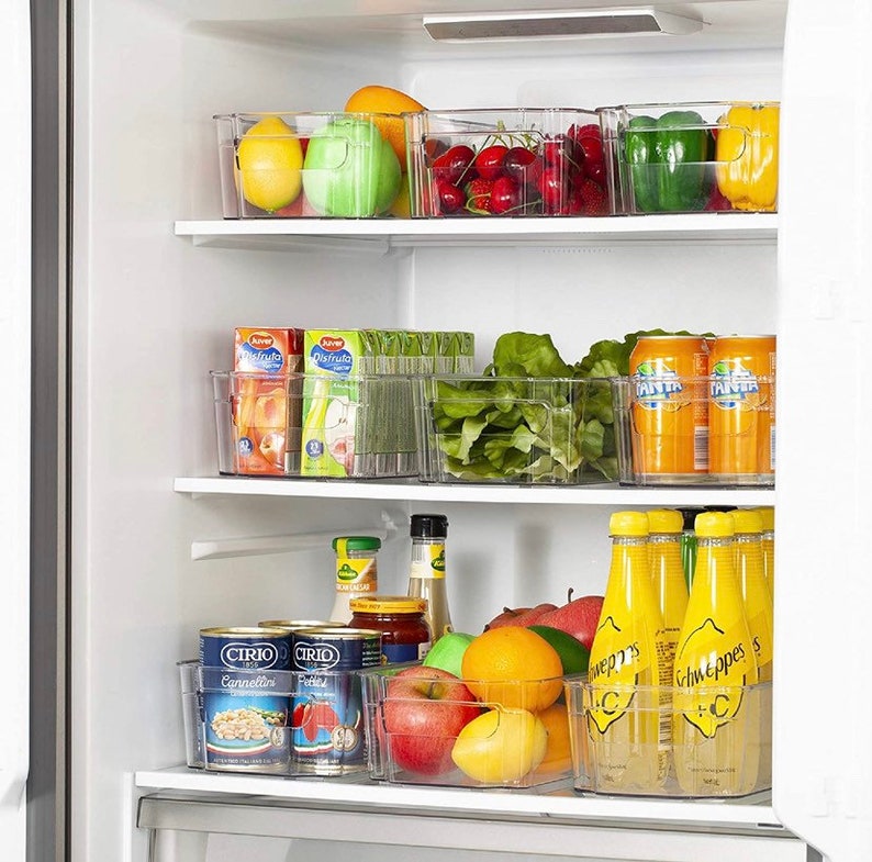 May include: A refrigerator with clear plastic storage containers filled with various fruits, vegetables, and beverages. The containers are stacked on three shelves. The top shelf contains red cherries, green bell peppers, and yellow bell peppers. The middle shelf contains green lettuce, orange juice boxes, and cans of orange soda. The bottom shelf contains canned goods, oranges, lemons, apples, and bottles of yellow lemonade.