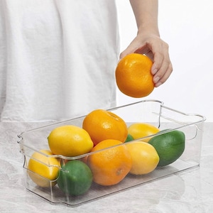 May include: A clear plastic refrigerator storage bin filled with oranges and limes. The bin has a handle on each side and is designed to stack.