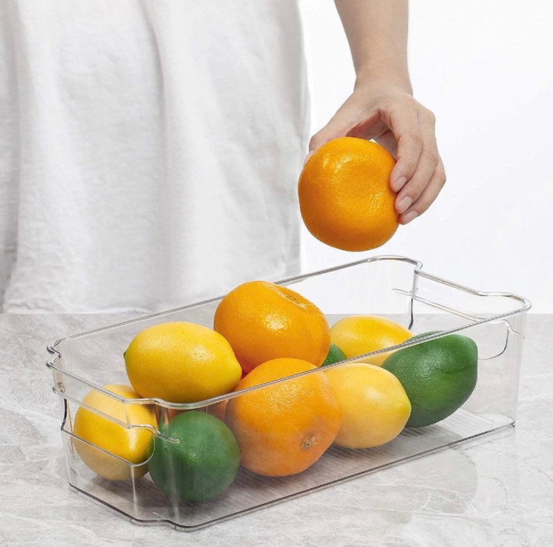 May include: A clear plastic refrigerator storage bin filled with oranges and limes. The bin has a handle on each side and is designed to stack.