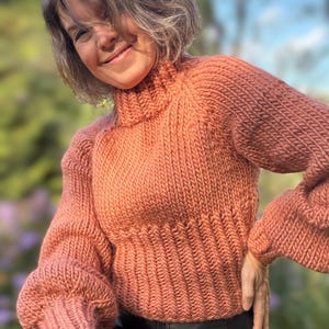 May include: A close-up of a knitted sweater in a warm peach color. The sweater has a high neck and a relaxed fit with puffed sleeves.
