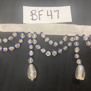May include: A decorative trim featuring clear, faceted beads with blue accents, suspended from a white satin ribbon. Two strands of beads hang in a scalloped pattern, with teardrop-shaped pendants. The tag reads "BF47".