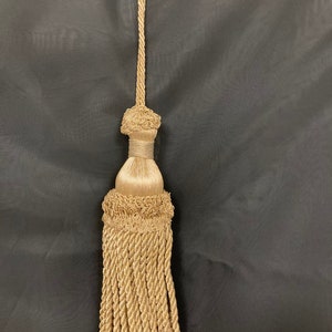 May include: A gold tassel with a long, twisted cord. The tassel has a decorative knot at the top and a fringe at the bottom.