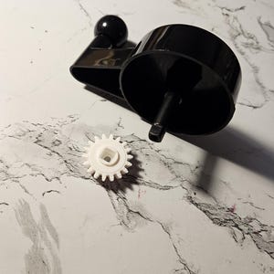 May include: A black plastic hand crank with a white plastic gear. The crank has a round knob on the end of the handle.