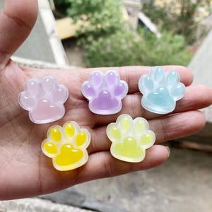 Puede incluir: Cinco dijes en forma de huella de pata en tonos rosa, morado, amarillo y azul. Los dijes son translúcidos y parecen estar hechos de resina o un material similar. Las huellas de pata tienen un diseño lindo y caprichoso.