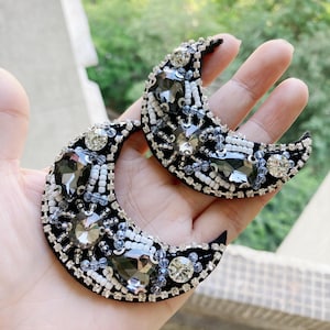 May include: Two black crescent moon shaped fabric patches with silver and white beads and rhinestones.