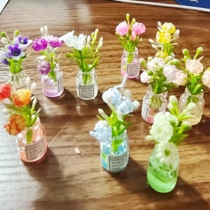 May include: A collection of miniature flower arrangements in small glass bottles. The flowers are various colors, including purple, pink, white, yellow, orange, and blue. Each bottle has a label with text.