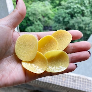 Może przedstawiać: Pięć żółtych chipsów ziemniaczanych na dłoni. Chipsy są okrągłe i mają fakturowaną powierzchnię.