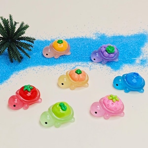 May include: A collection of seven colorful, translucent turtle figurines. Each turtle has a different fruit or vegetable design on its shell, including a tomato, strawberry, pumpkin, and more. The turtles are arranged on a white surface with blue sand.