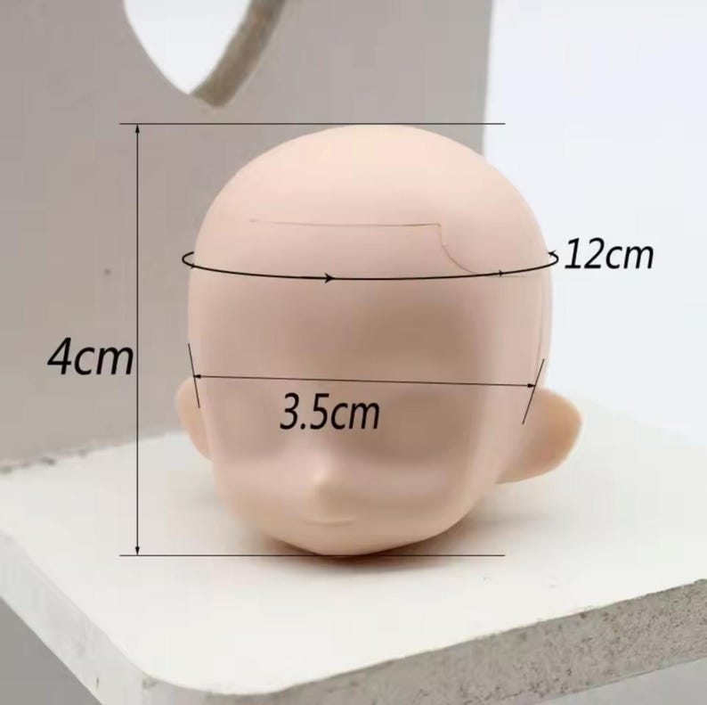 May include: A blank doll head made of a smooth, light-colored material. The head is 12 cm wide and 4 cm tall. The head has a 3.5 cm wide opening for the neck.