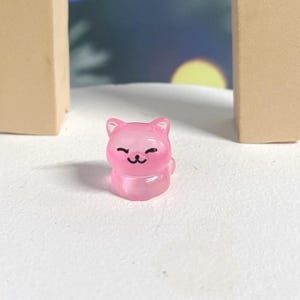 May include: A small, pink, translucent cat figurine with a smiling face. The figurine is made of resin and has a glossy finish.