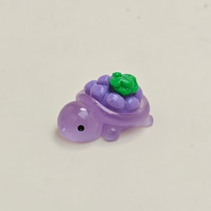 May include: A small, purple turtle figurine with a cluster of purple grapes and a green leaf on its back. The turtle has a black eye and is made of a translucent material.