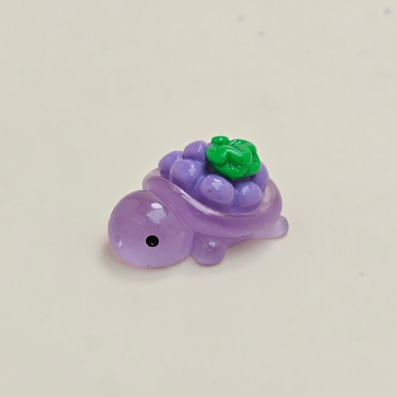 May include: A small, purple turtle figurine with a cluster of purple grapes and a green leaf on its back. The turtle has a black eye and is made of a translucent material.