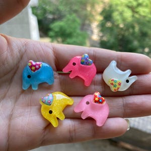 Könnte beinhalten: Fünf bunte Plastik-Elefantenfiguren mit floralen Herzdesigns. Die Elefanten sind in den Farben Blau, Rosa, Gelb und Weiß.