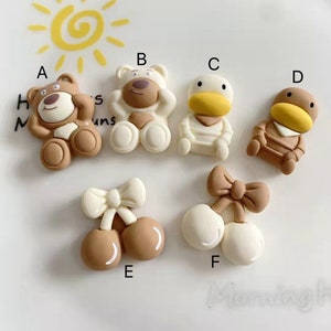 Pode incluir: Conjunto de seis enfeites planos de resina em várias formas de animais e alimentos. Os enfeites são em tons de marrom, branco e amarelo. Os animais incluem um urso, um pato e um pato vestindo uma camisa listrada. Os alimentos incluem um par de cerejas com um laço e um par de cerejas com um laço.