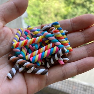 May include: A pile of colorful, twisted, candy cane-shaped polymer clay charms. The charms are in various colors, including pink, blue, yellow, white, brown, and purple.