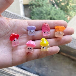 May include: Six miniature mushroom figurines with different colors and facial expressions. The mushrooms are made of plastic and have a glossy finish.