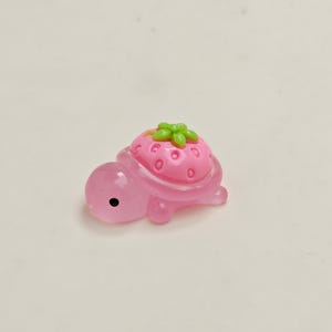 May include: A small, pink turtle figurine with a strawberry shell. The strawberry is pink with green leaves and small, red seed details. The turtle has a black eye and is made of a translucent material.
