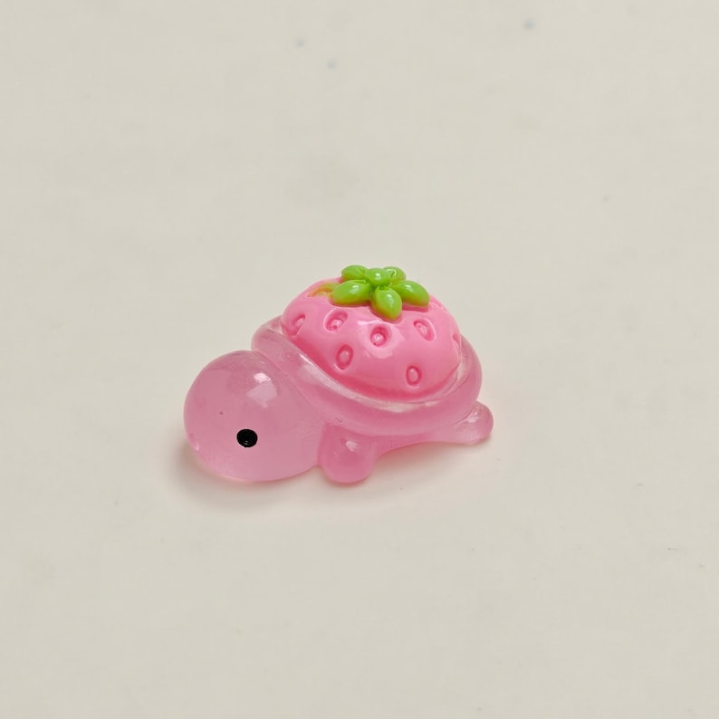 May include: A small, pink turtle figurine with a strawberry shell. The strawberry is pink with green leaves and small, red seed details. The turtle has a black eye and is made of a translucent material.