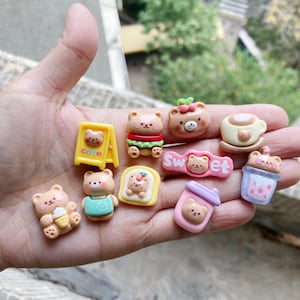 Puede incluir: Una colección de 10 encantos de resina coloridos con osos de dibujos animados lindos en varias poses. Los encantos incluyen un letrero de café, un sándwich, una taza de café, un té de boba, un letrero de dulce, un oso sosteniendo un cono de helado, un oso sosteniendo una bebida, un oso sosteniendo una hamburguesa y un oso sosteniendo una rebanada de pan. Los encantos son perfectos para agregar un toque de fantasía a tus manualidades.