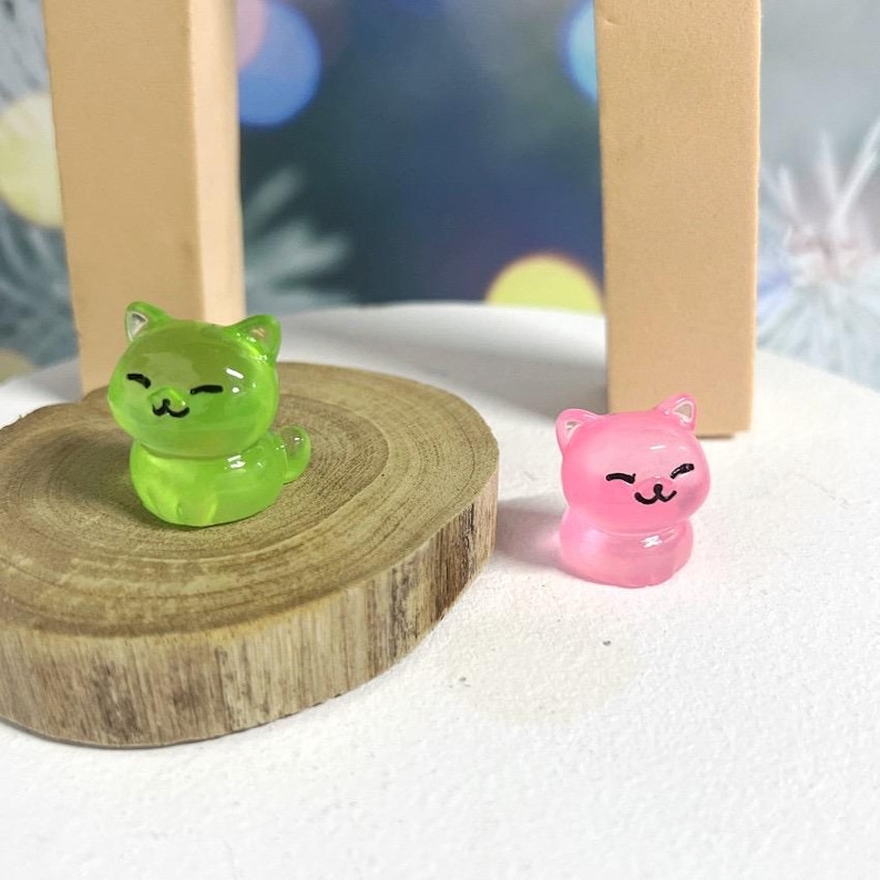 May include: Two small, clear plastic figurines of cats. The cat on the left is green and the cat on the right is pink.