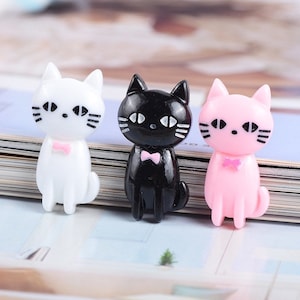 May include: Three small plastic cat figurines. The cats are white, black, and pink. Each cat has a pink bow tie and black eyes.