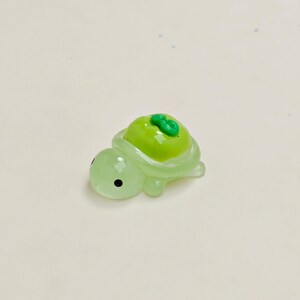 May include: A small, translucent green turtle figurine with a bright green apple on its back. The turtle has black eyes and is made of a glossy material. The apple has a small green stem.