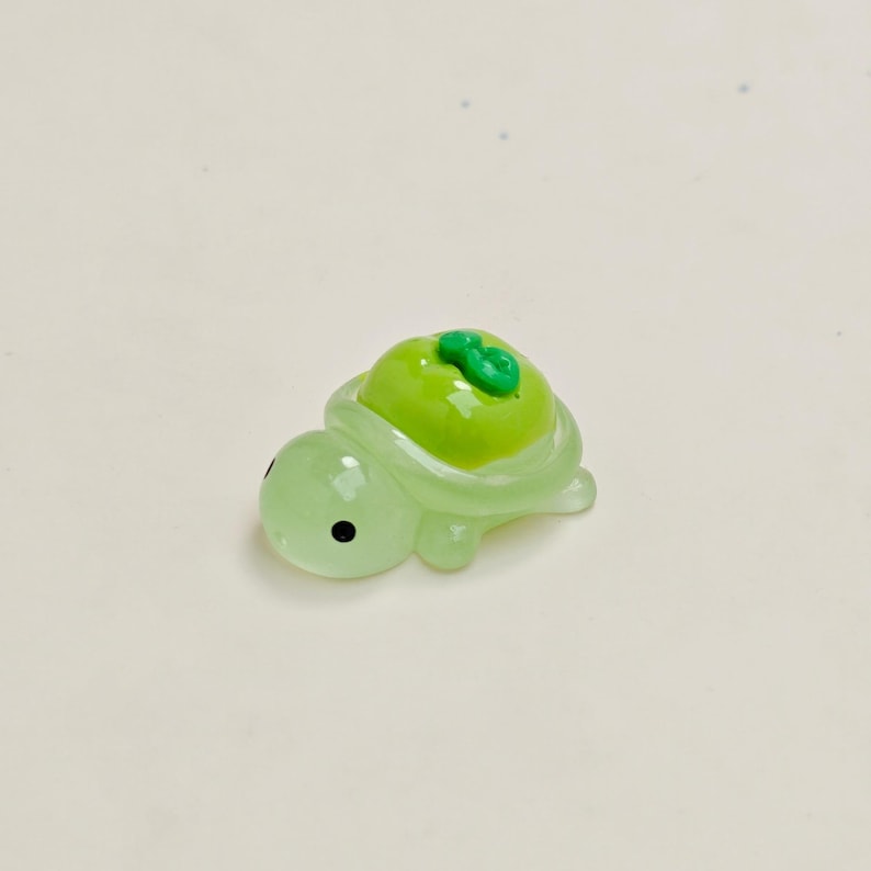 May include: A small, translucent green turtle figurine with a bright green apple on its back. The turtle has black eyes and is made of a glossy material. The apple has a small green stem.