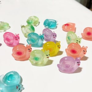 以下が含まれることがあります： 半透明のプラスチックでできたカラフルなカタツムリの置物のコレクション。カタツムリはピンク、ブルー、グリーン、イエロー、パープルなど、さまざまな色をしています。各カタツムリは、笑顔が描かれた顔をしています。