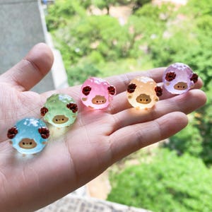 Op de afbeelding: Een hand die zes kleine, kleurrijke schapenbeeldjes vasthoudt. Elk schaap heeft een rond lichaam en een klein gezicht met twee zwarte stippen als ogen. De schapen zijn in tinten blauw, groen, roze, oranje en paars. De schapen hebben bruine hoorns en zijn doorschijnend.