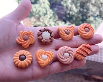 May include: A collection of eight miniature cookies in various shapes and colors, including heart, flower, and swirl designs. The cookies are made of polymer clay and are perfect for crafting or decorating.