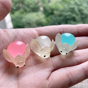 Pode incluir: Três pequenos brinquedos em forma de inseto, translúcidos, com corpo redondo e olhos pretos. Os brinquedos são feitos de um material plástico transparente e são cheios de líquido colorido. As cores do líquido são rosa, branco e azul.
