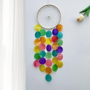 Puede incluir: Un atrapasueños arcoíris con una red de macramé blanca y un anillo de madera. El atrapasueños está decorado con cuentas redondas de colores que cuelgan de la parte inferior.