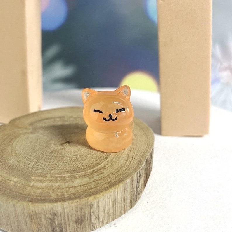 May include: A small, orange resin figurine of a cat with a winking face. The figurine is sitting on a wooden surface.