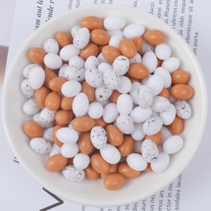 Puede incluir: Un tazón blanco lleno de una mezcla de huevos decorativos ovalados de color marrón y blanco moteado. Los huevos están dispuestos en un patrón aleatorio.