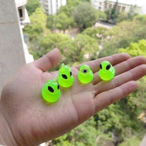 May include: Four small, glowing green alien figures with black eyes are held in a hand. The figures have different designs, with one having a single eye. The background is blurred, showing green trees and buildings.