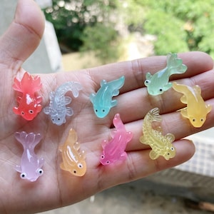 May include: Nine miniature goldfish figurines in various colours, including red, blue, green, yellow, pink, and purple. The translucent fish are held in a hand, highlighting their intricate details. Each fish is a unique design.