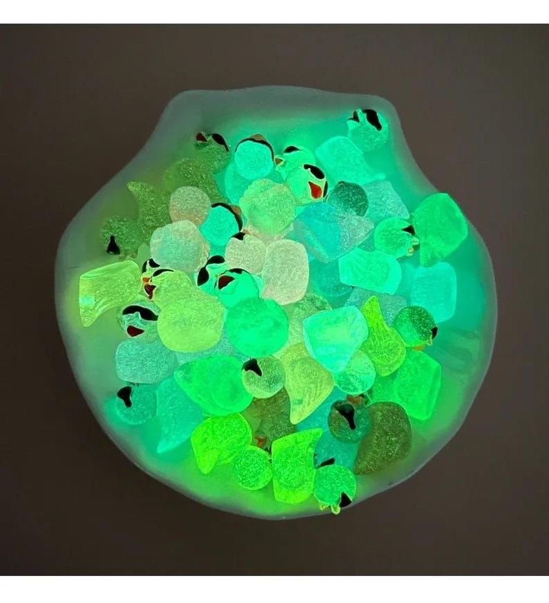 May include: A white seashell filled with glowing green, blue, and yellow glass pebbles. The pebbles are arranged in a random pattern and are illuminated by a black light.
