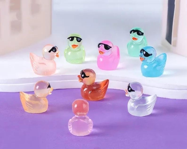 May include: A set of nine colorful rubber duck figurines with sunglasses. The ducks are made of clear plastic and have different colors, including pink, blue, green, and yellow. Each duck has a pair of black sunglasses.
