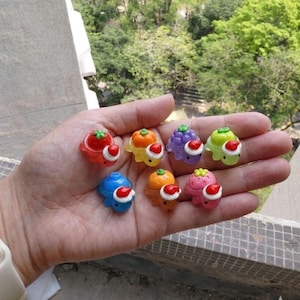 Pode incluir: Uma mão segurando uma coleção de pequenas figuras coloridas em forma de fruta. Cada figura é de uma cor diferente, incluindo vermelho, laranja, roxo e verde, e tem um gorro de Papai Noel branco e vermelho. As figuras são feitas de um material translúcido.