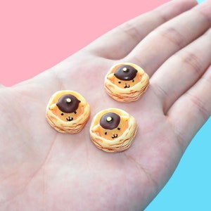 Puede incluir: Tres figuritas de panqueques en miniatura de cerámica con cobertura de chocolate marrón y una linda carita de dibujos animados. Los panqueques son amarillos y tienen una textura en capas.