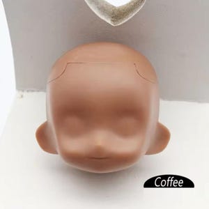May include: A close-up of a doll head with a coffee-colored skin tone. The head is made of a smooth, plastic material and has a slightly open mouth. The doll head is sitting on a white surface.