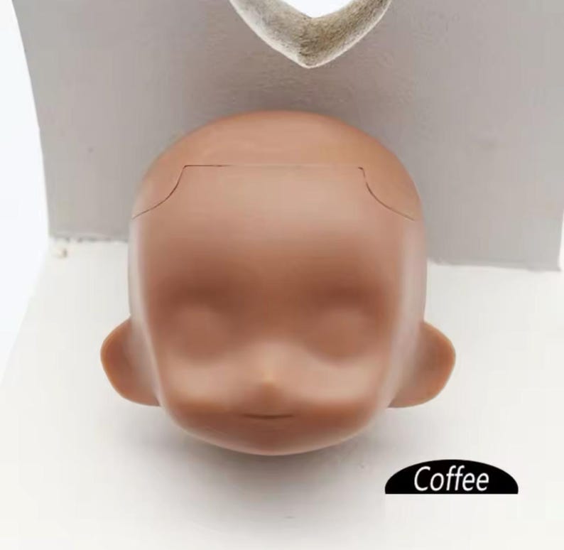 May include: A close-up of a doll head with a coffee-colored skin tone. The head is made of a smooth, plastic material and has a slightly open mouth. The doll head is sitting on a white surface.