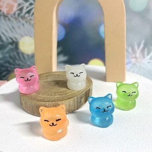 May include: Five small, colourful, resin cat figurines. The cats are all in a sitting position with their paws tucked under them. They are all smiling and have black eyes. The cats are in the following colours: pink, white, green, blue, and orange.