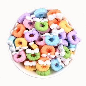 Può includere: Una ciotola piena di ciondoli colorati a forma di ciambella in miniatura con fiocchi bianchi. I ciondoli sono in tonalità di rosa, blu, verde, arancione e viola.