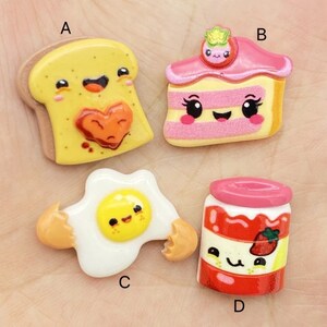 Op de afbeelding: Vier kleurrijke, cartoonachtige voedselcharms, waaronder een stuk taart, een gebakken ei, een pot jam en een snee toast. Elke charme heeft een schattig gezicht en is gemaakt van hars.