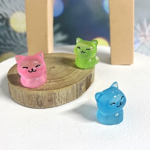 May include: Three small, clear plastic figurines of cats in pink, green and blue. Each cat has a smiling face with closed eyes and a small tail.