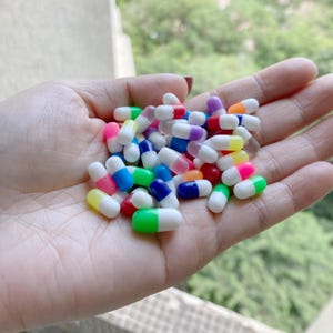 Può includere: Una mano che tiene un grande mucchio di capsule di plastica colorate. Le capsule sono bianche con una varietà di colori alle estremità, tra cui rosso, blu, verde, giallo e viola.
