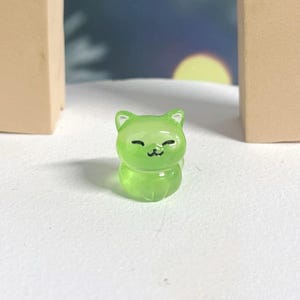 May include: A small, green, glass cat figurine with a cute, smiling face. The figurine is sitting on a white surface.