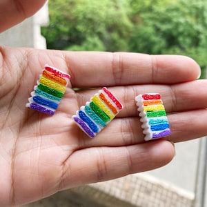 May include: Three miniature rainbow layer cakes with white frosting. Each cake has six layers of frosting in the colors of the rainbow.