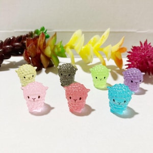 May include: A set of eight miniature sheep figurines in various pastel colors, including pink, light blue, green, purple, yellow, and gray. The sheep are made of plastic and have a textured surface.